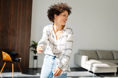 Modern bir dairede gündelik kıyafetlerle dans eden mutlu genç bir kadın.