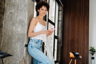 Gülümseyen kıvırcık kadın modern bir dairede pencerede duruyor.