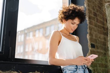 Gülümseyen kıvırcık kadın modern bir dairede pencerede duruyor.