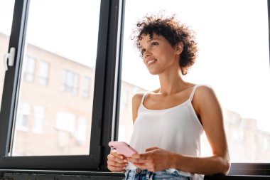 Gülümseyen kıvırcık kadın modern bir dairede pencerede dikiliyor.