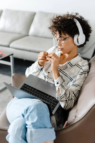 坐在家里扶手椅上使用笔记本电脑的年轻卷曲女人 — 图库照片
