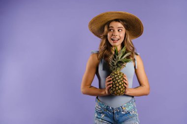 Hasır şapkalı, gülümseyen ve elinde mor arka planda izole edilmiş ananas tutan genç bir kadın.