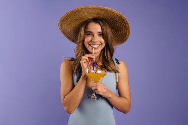 Mayo ve hasır şapkalı genç beyaz kadın mor arka planda izole edilmiş kokteyl içiyor.