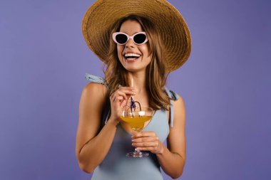 Güneş gözlüklü ve hasır şapkalı genç beyaz kadın mor arka planda izole edilmiş kokteyl içiyor.