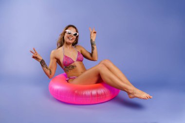 Mayo giymiş, gülümseyen ve kauçuk halkada oturan, mor arka planda soyutlanmış genç bir kadın.