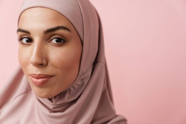 Pembe tesettürdeki Müslüman kadının pembe stüdyoda dikilirken kameraya bakışının yakın çekimi.
