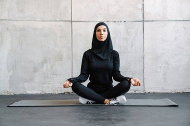 İçeride fitness minderinde otururken meditasyon yapan tesettürlü genç Müslüman kadın.