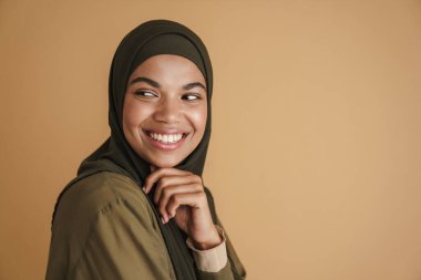tesettürlü siyah müslüman kadın gülümsüyor ve bej arka planda soyutlanmış bir kenara bakıyor.