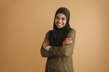 Tesettürlü siyah Müslüman kadın gülümsüyor ve bej arka planda izole edilmiş kameraya bakıyor.