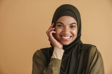 Tesettürlü siyah Müslüman kadın gülümsüyor ve bej arka planda izole edilmiş kameraya bakıyor.