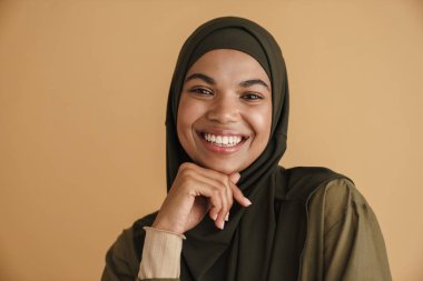 Tesettürlü siyah Müslüman kadın gülümsüyor ve bej arka planda izole edilmiş kameraya bakıyor.