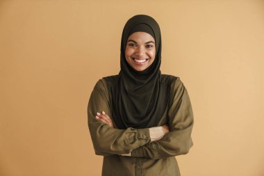 Tesettürlü siyah Müslüman kadın gülümsüyor ve bej arka planda izole edilmiş kameraya bakıyor.