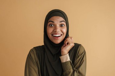 Tesettürlü siyah Müslüman kadın gülümsüyor ve bej arka planda izole edilmiş kameraya bakıyor.