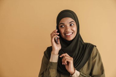 Bej arka planda izole edilmiş, tesettürlü, gülümseyen ve cep telefonuyla konuşan siyah Müslüman kadın.