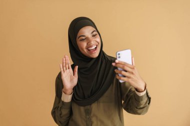 Bej arka planda izole edilmiş cep telefonundan selfie çeken tesettürlü siyah Müslüman kadın.