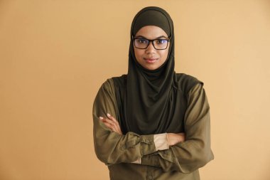 Siyah Müslüman kadın tesettürde poz veriyor ve bej arka planda izole kameraya bakıyor.