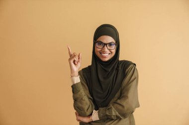 Tesettürlü siyah Müslüman kadın gülümsüyor ve bej arka planda tek başına parmağını yukarı kaldırıyor.