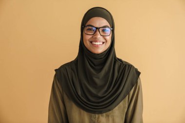 Tesettürlü siyah Müslüman kadın gülümsüyor ve bej arka planda izole edilmiş kameraya bakıyor.