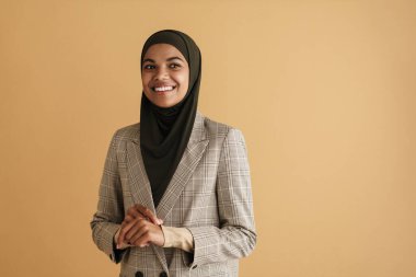 Bej arka planda izole edilmiş kameraya poz verirken tesettürlü siyah Müslüman kadın gülümsüyor
