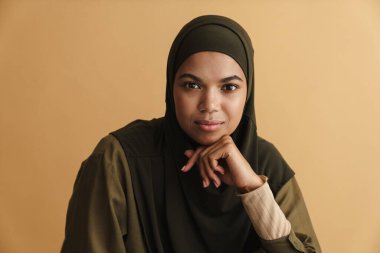 Siyah Müslüman kadın tesettürde poz veriyor ve bej arka planda izole kameraya bakıyor.