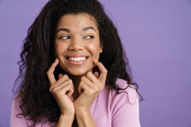 Genç siyah kadın gülümsüyor ve mor arka planda izole edilmiş yanaklarını işaret ediyor.