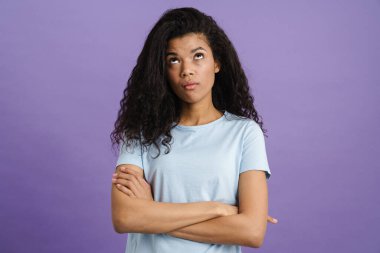 Kolları çaprazlanmış kıvırcık poz veren, kafası karışmış genç bir kadın mor arka planda izole edilmiş.