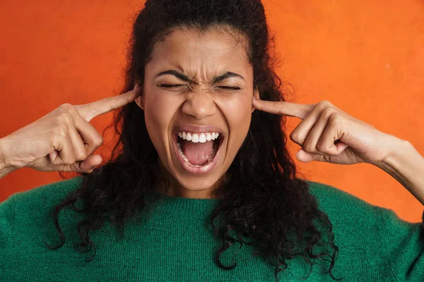 Frowning young african woman in casual wear standing over orange background, screaming, blocking ears