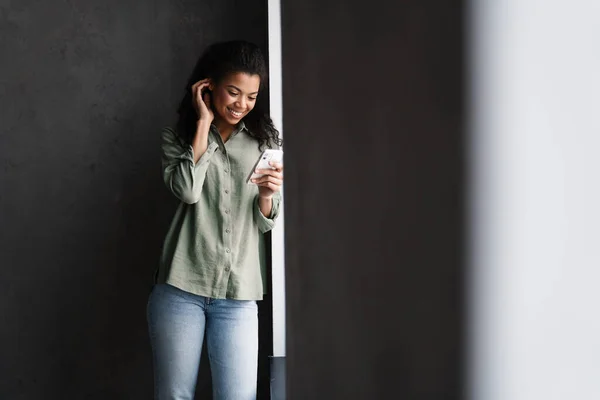 Musta Nuori Nainen Käyttää Matkapuhelinta Nojaten Seinään Sisätiloissa — kuvapankkivalokuva