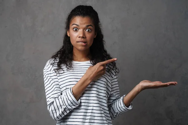 Shocked african casual brunette woman standing over gray wall background, presenting copy space