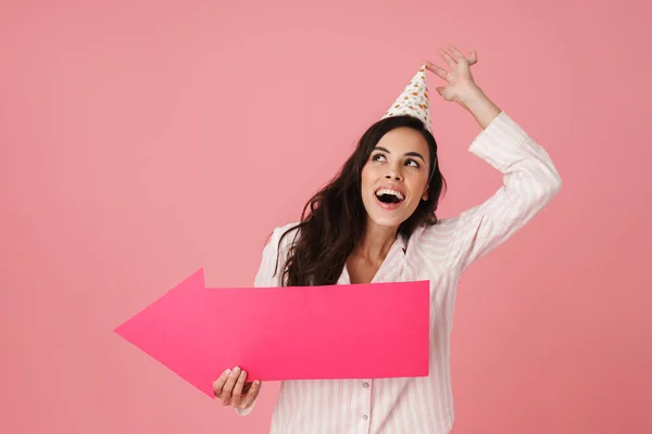 穿着派对圆锥的快乐女人一边笑一边拿着箭头标牌站在粉色背景上 — 图库照片