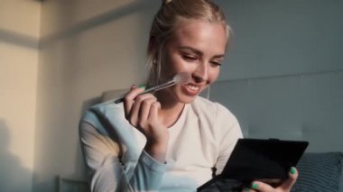 Gülümseyen bir kadın dairenin içinde otururken yanaklarına allık sürüyor.