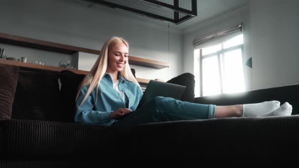 一位面带微笑的金发女商人正坐在公寓里的沙发上 用她的笔记本电脑上的视频连接工作 — 图库视频影像