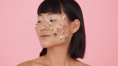 Yüzünde renkli çiçeklerle kameraya poz veren muhteşem Asyalı bir kadın stüdyoda pembe bir arka planın başında duruyor.