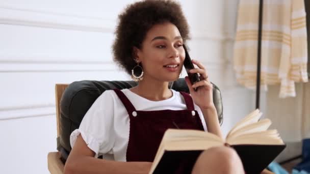 一个漂亮的非洲裔美国女人在屋里坐在椅子上一边看书一边用手机聊天的特写镜头 — 图库视频影像