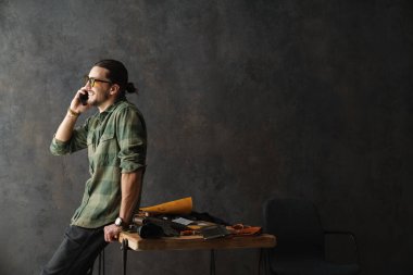 Sakallı, gülümseyen bir zanaatkar atölyede deriyle çalışırken cep telefonuyla konuşuyor.