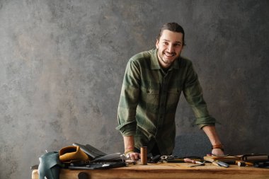 Gülümseyen orta yaşlı deri ustası atölyesinde çalışıyor, masada duruyor.