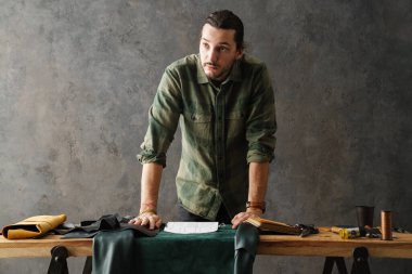 Sakallı, odaklı bir zanaatkar atölyede masada dururken deriyle çalışıyor.