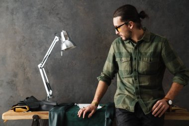 Beyaz sakallı bir zanaatkar atölyede masa başında çalışırken deriyle çalışıyor.