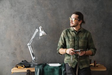 Sakallı beyaz zanaatkar atölyesinde deri debriyajla dikiliyor.