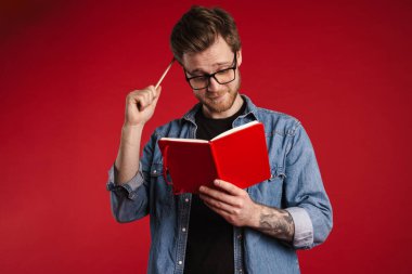 Düşünceli düşünceli genç adam kırmızı duvarın arkasında durmuş, kitap okuyor, elinde kalem tutuyor.