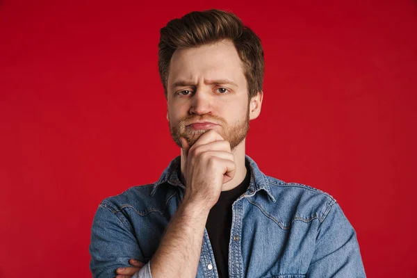 Pensive young white man in casual clothes standing isolated over red, thinking