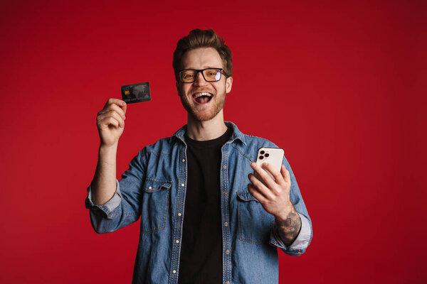 Happy smiling young man in casual clothes standing over red wall background, holding mobile phone and credit card, shopping, celebrating success