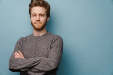 Beyaz kızıl adam kameraya bakarken kolları çaprazlanmış bir şekilde mavi arka planda duruyor.