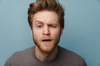 Beyaz, sakallı, suratını buruşturan ve mavi arka planda izole edilmiş kameraya bakan kızıl adam.