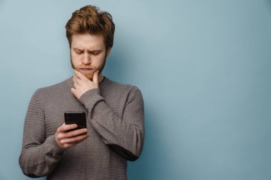 Kafası karışmış genç, düşünceli, sıradan bir adamın portresi. Gri arka planda duruyor. Elinde cep telefonu, çenesine dokunuyor.