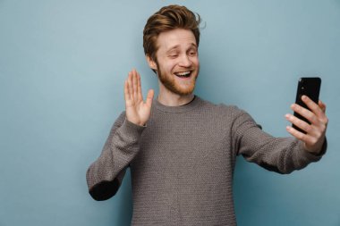 Beyaz kızıl adam mavi arka planda izole edilmiş cep telefonuyla selfie çekerken işaret veriyor.