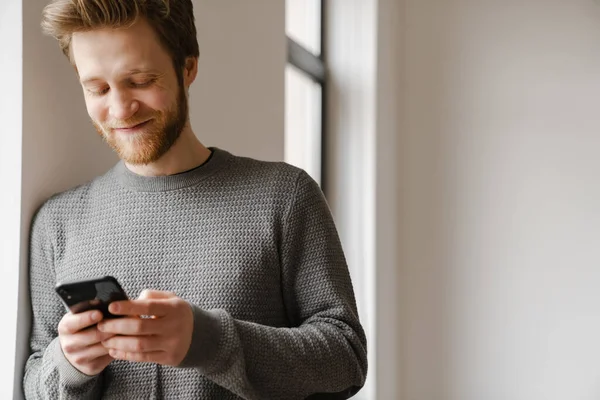 Ginger Young Man Using Mobile Phone While Leaning Wall Indoors — Stock Photo, Image