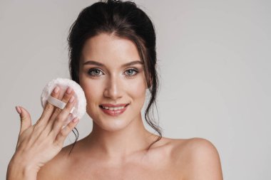 Kahverengi saçlı, gülümseyen beyaz bir kadın elinde pudra ponponu tutuyordu. Gri arka planda izole olmuştu.