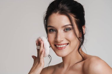 Kahverengi saçlı, gülümseyen beyaz bir kadın elinde pudra ponponu tutuyordu. Gri arka planda izole olmuştu.