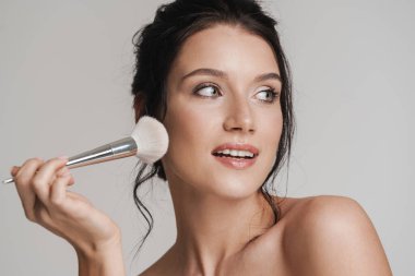 Kahverengi saçlı, gülümseyen beyaz kadın elinde makyaj fırçası, gri arka planda izole edilmiş.